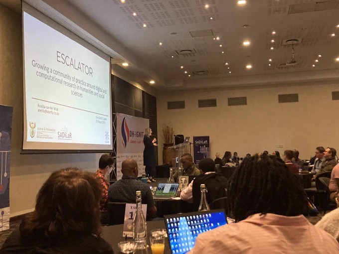 A conference room with round tables and attendees sitting in groups around those tables. Almost all of them have their backs to the camera, facing the front. At the front is a screen on which is projected "ESCALATOR: Growing community of practice around digital and computational research in humanities and social sciences" and on the right side of the screen is a banner with DH-IGNITE printed on it. Standing in front of the screen is Anelda van der Walt, a Consultant & Digital Literacy Trainer with the ESCALATOR programme, as well as a Carpentries Instructor, telling the participants more about ESCALATOR. 