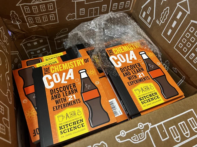An open cardboard box filled with copies of a book called The Chemistry of Cola