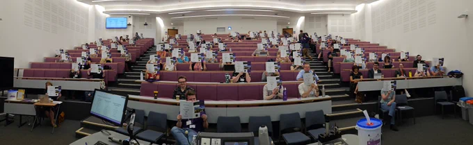 Attendees of ViCEPHEC 2023 sitting in the main lecture theatre holding up conference leaflets to their faces.