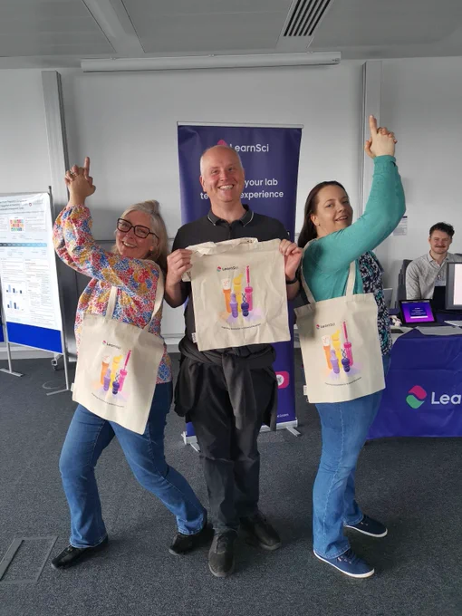 Three people in a line holding LearnSci tote bags, the person in the centre facing the camera and the people on the sides standing with their backs to the person in the center, with their hands in the air making a gun-shaped gesture.