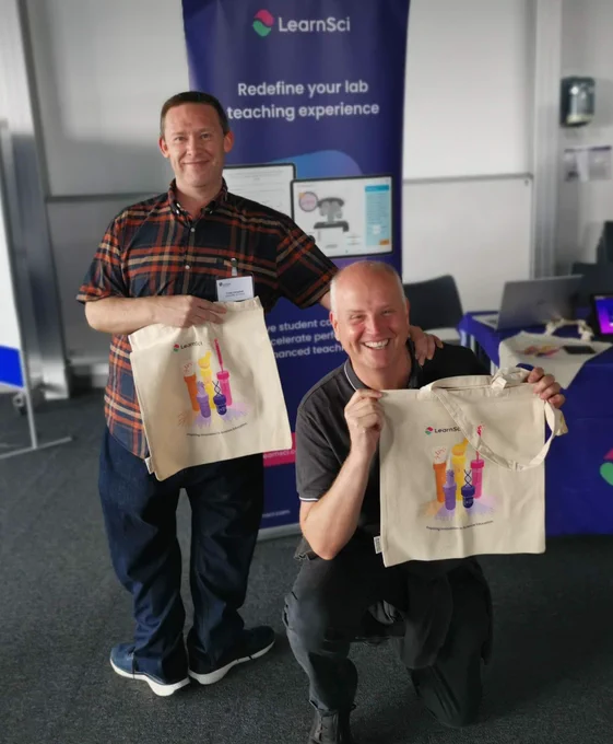 Two people, smiling, holding LearnSci tote bags, one standing, one kneeling on the ground.