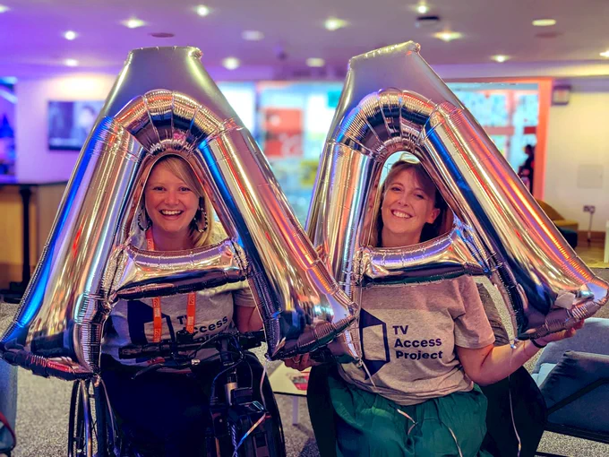 Bryony and Ally each holding  a silver A helium balloon, poking their faces through and smiling
