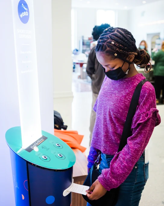 A girl receiving a short story or poem from the Short Story Dispenser at Planet Word. The machine has three buttons and a backlit sign with instructions on top, and it dispenses stories and poems on a strip of paper.