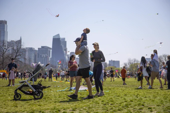 Image shows a little boy on a man’s shoulders in Zilker Park on a sunny day. The little boy is looking up at the sky seeing the colorful kites that are flying around. Lots of people and families are around them. Some are holding kites to prepare them to go in the sky while others are also carrying kids on their shoulders. Photo by Julius Shieht. 