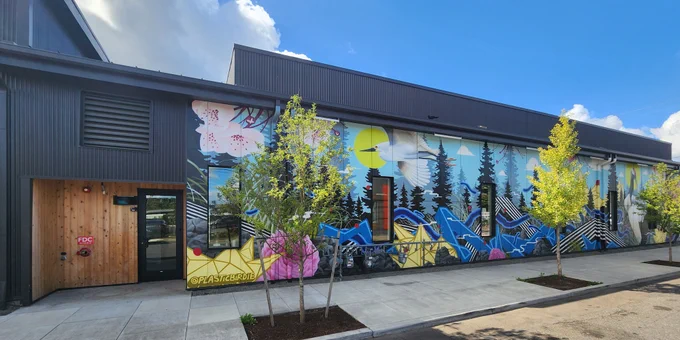 View of the side of the CORE PDX building which includes the side-entry door on the left and colorful mural with flowers, trees, and varying shapes on the right appears behind a few newly planted trees sprinkled along the sidewalk next to the building.