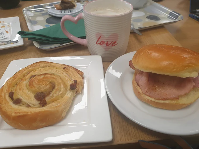 A raisin pastry, bacon roll and cup of coffee.