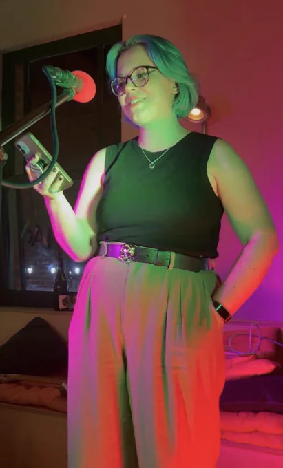 A white femme person with blue hair on stage reading from their phone, cast in red and green light