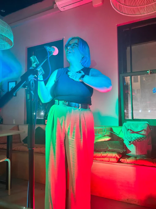 A white femme person with blue hair on stage reading from their phone, cast in red, blue and green light. Their hand is on their chest and they are smiling.