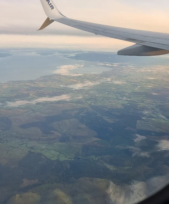 Image shows green fields and early morning fog out an airplane window with the plane wing in sight