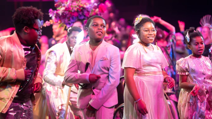 A group of children on stage in white and silver outfits. They dance and sing and are lit with a pink light.