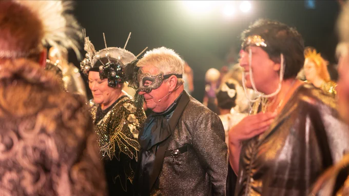 A group of people dance in black and gold embellished outfits and wear masks and headdresses.
