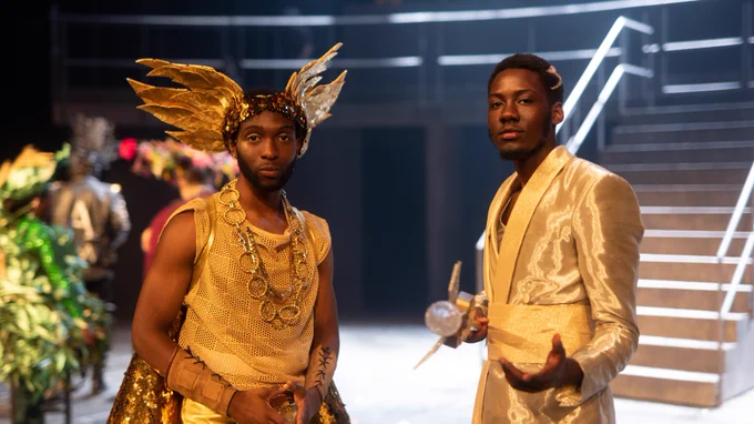 Two young men in gold outfits and headdresses look straight at the camera. Behind them is a staircase up to a balcony.