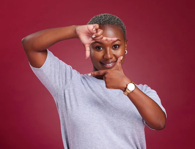A woman holding up her hands in a "L" shape as a camera against a maroon background.