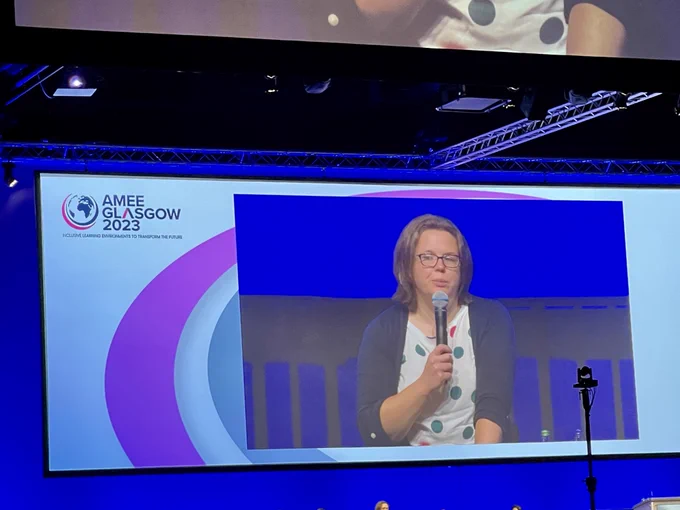 Screen of key note. On left, logo for AMEE Glasgow 2023, white female in multi-colored blouse and glasses holding microphone. 