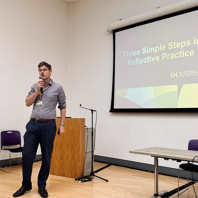 Chris stands at the top of a room speaking to the audience with a slide behind him reading “Three Simple Steps to Reflective Practice”