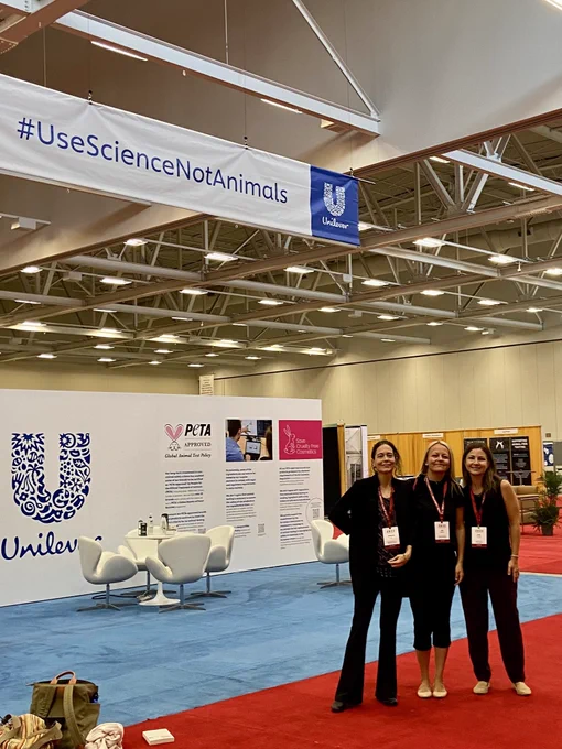 Three women stand together in a conference exhibit hall. A team united to make science better for humans and non-humans. Their smiles beam under a sign (sponsored by Unilever) reading, “#UseScienceNotAnimals.”