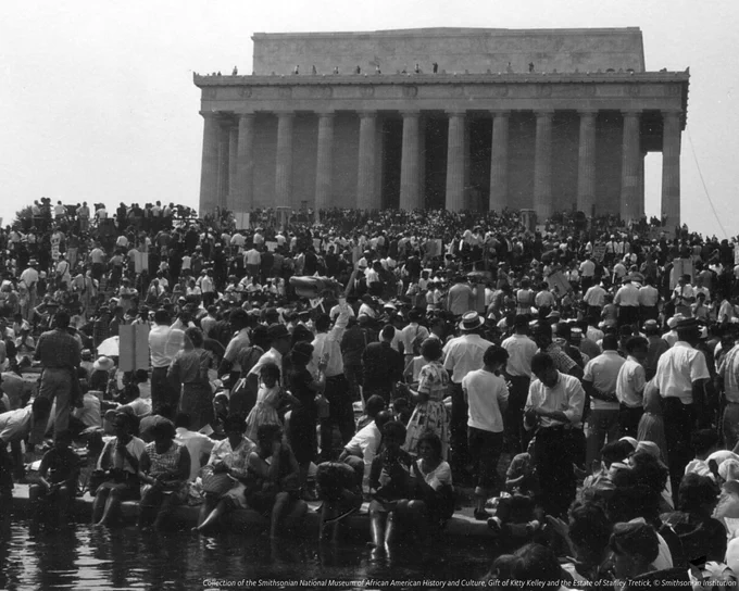  A black and white photo of the March on Washington crowd standing in front of the Lincoln Memorial on August 28, 1963. Some participants are crowded around the edge of the Reflecting Pool. 
