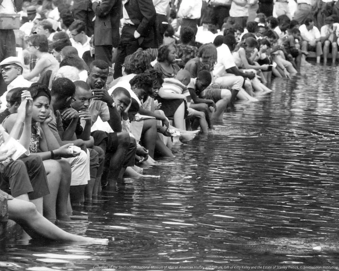 A black and white photo of March on Washington participants sitting on the end of the Reflecting Pool with their feet submerged on August 28, 1963. The dark water has ripples across the surface. Behind the first row of people sitting with their feet submerged are a crowd of others, mostly sitting facing the opposite direction. 

 
