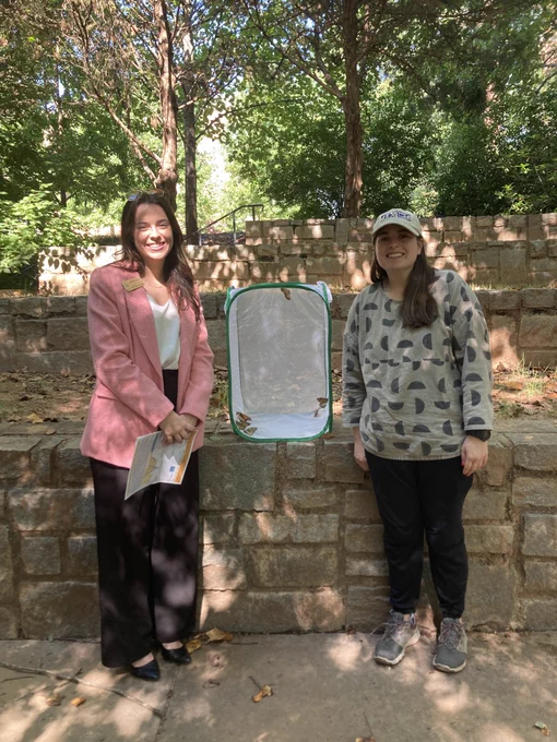 A congressional staffer and a graduate student are posed facing the camera. They are outside. There is a netted box between them containing monarch butterflies used for research