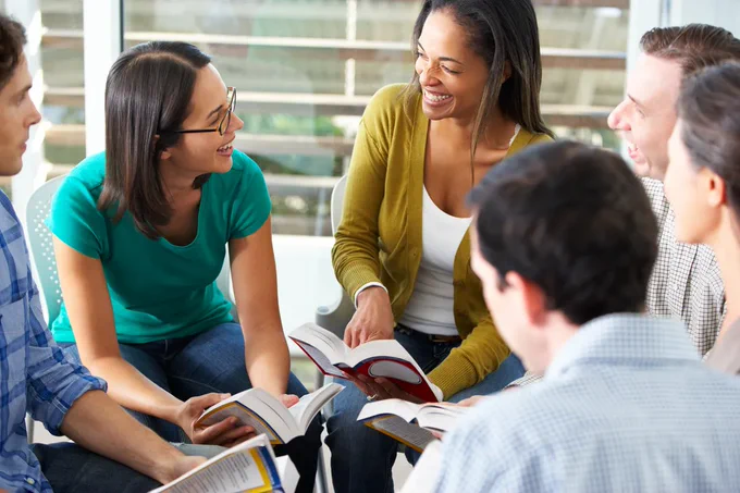 A group of six people sitting on chairs in a circle reading books and smiling at each other. Though, upon closer inspection these books look like dictionaries.