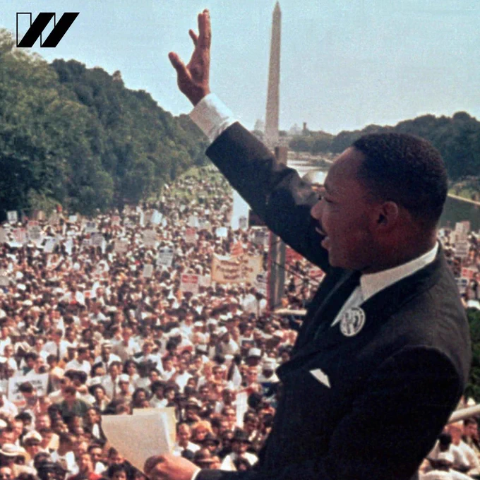 Image description: An image of Martin Luther King Jr. with his hand raised in front of the crowd listening to his speech at the March on Washington. There is a black “w” U S O W logo in the top left corner.