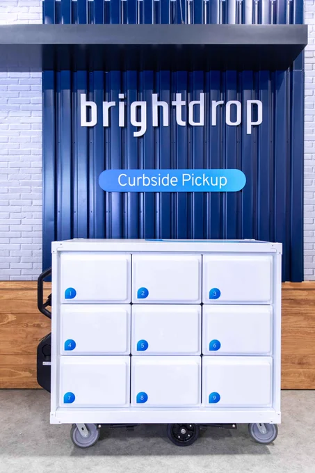 A BrightDrop Trace Grocery sits in front of a blue wall that reads "BrightDrop" and "Curbside Pickup"