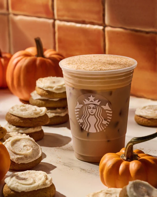 A Starbucks Iced Pumpkin Cream Chai Tea Latte rests in an orange-tiled kitchen next to mini pumpkins and chai frosted cookies.