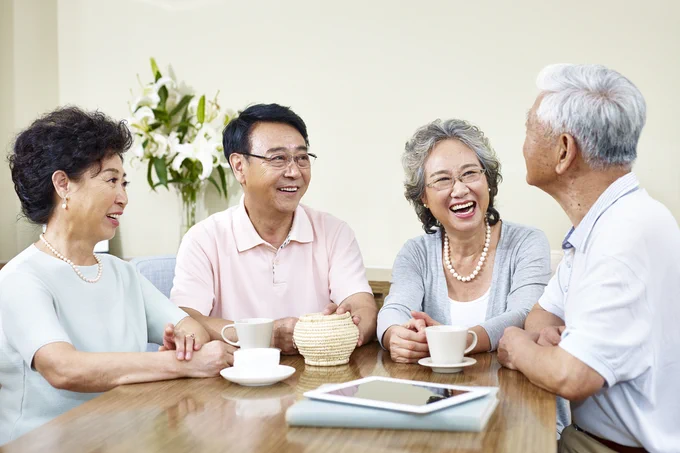 A group of four Korean seniors sitting around a table with coffee cups. There's an iPad and a book on the top of the table. They're all conversing with and smiling at each other.