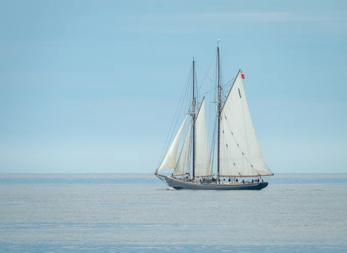Bluenose II with the 4 lower sails set, with a baby blue sky and light blue water, sailing to the left.