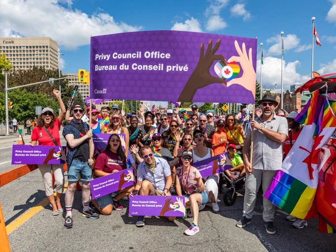 A group of employees from the Privy Council Office posing for a photo together alongside Clerk Hannaford with a purple banner and signage that reads: Privy Council Office. Bureau du Conseil privé.