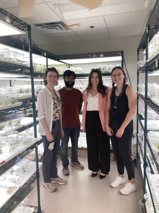 Three graduate students are standing with one congressional staffer. They are surrounded by shelves that hold plants that used for research