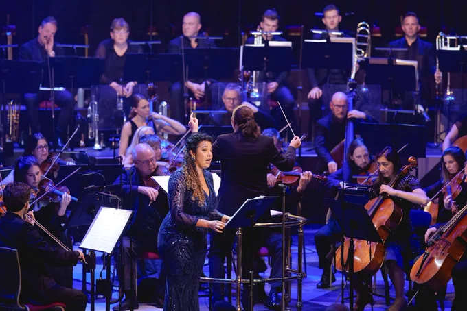Mezzo soprano Felicity Buckland sings on stage holding the music stand in front of her. The conductor behind her has her back to us with her hands raised at the orchestra.