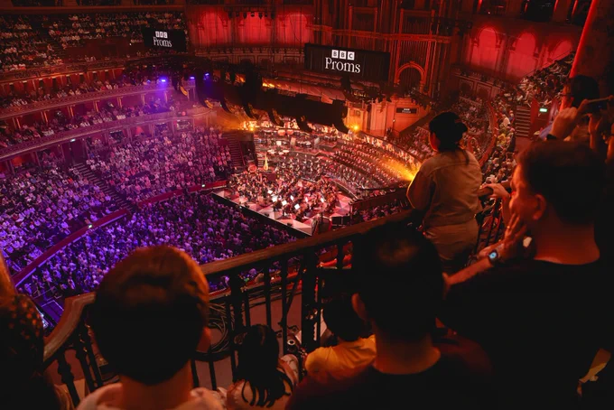 The shot looks down from behind the audience in the gallery on the orchestra below on the stage. You can also see the audience in the distance below.