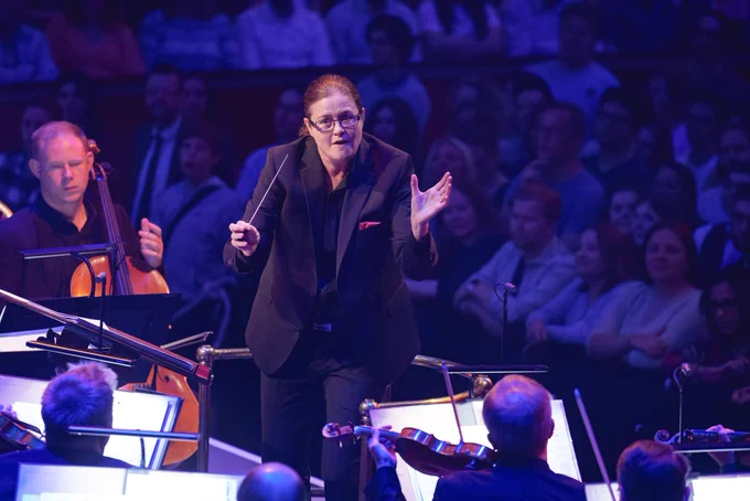 Conductor Anna-Maria Helsing holds her baton in one hand and has her other hand open in a gesture to the musicians in front of her. Prommers stand behind watching on.