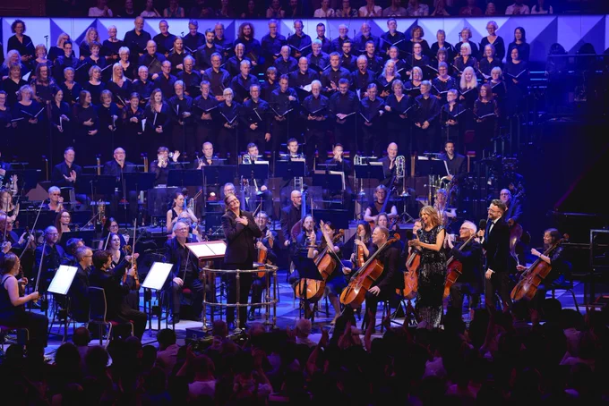 Conductor Anna-Maria Helsing stands on a podium with both hands on her heart. Katie Derham looks at her smiling and clapping her hands. Ali Plumb also smiles. The musicians in the orchestra also smile and clap.