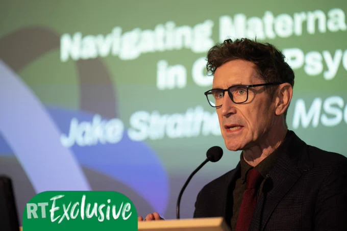 Paul McGann as Jake in Annika season 2 standing in front of a microphone on stage with a presentation bearing the character's name behind him