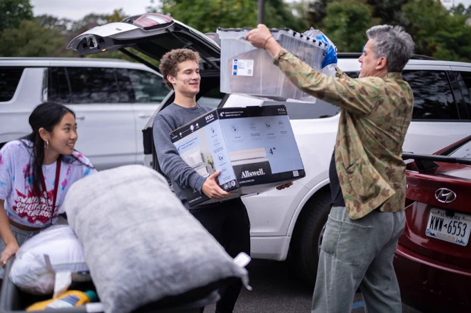 Three people standing by a car. They are holding boxes.