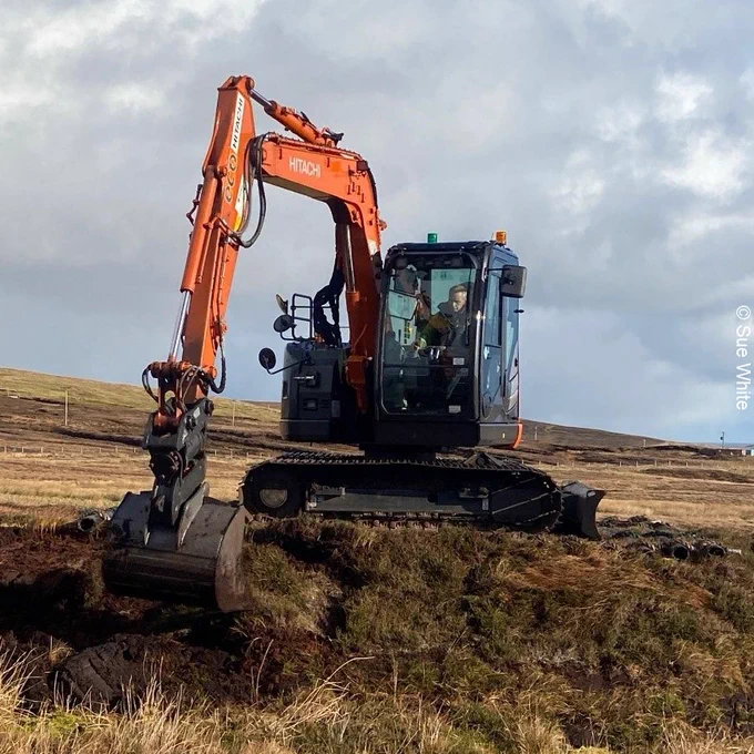 An operator in an orange excavator moves peatland vegetation as part of peatland restoration work.  Image credit: Sue White.