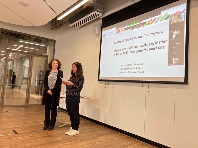 Two women standing in front of a big screen. One woman is reading from a piece of paper, the other is looking at the camera and smiling.