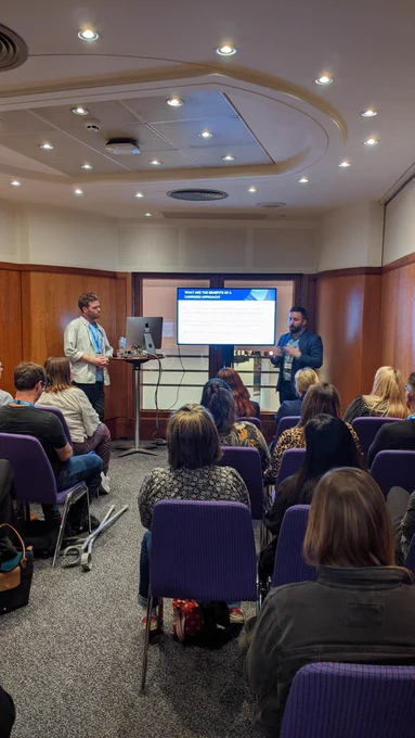 David and Tony standing at the front of a room with a PowerPoint presentation. Audience in rows in front of them. 