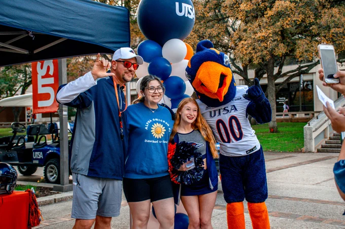 Coach Traylor, Rowdy, a cheerleader (Noelle Pratt) and a student smile at the camera and do the Roadrunner handsign. 