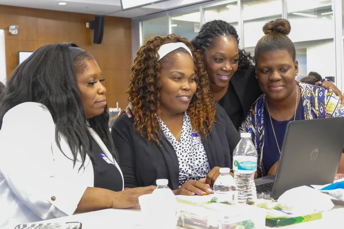 Four women smile as they look at a laptop monitor that is facing towards them. 