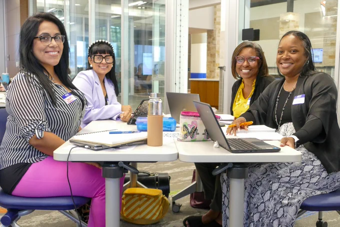 Four women sitting at a table smile for a photo. They have their laptops opened up ready to take notes. 