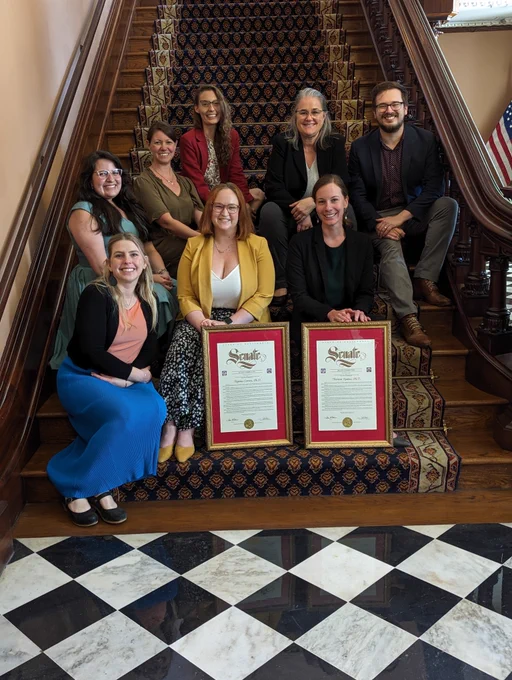A group of legislative staff sitting on the indoor stairs of the California State Capitol holding two framed Senate recognitions with visible black and white checkerboard floor.