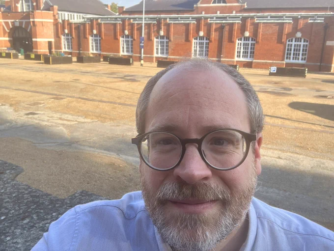 A selfie of a man with a greying beard in front of a long, red-brick building which seems to glow in the morning sunshine.