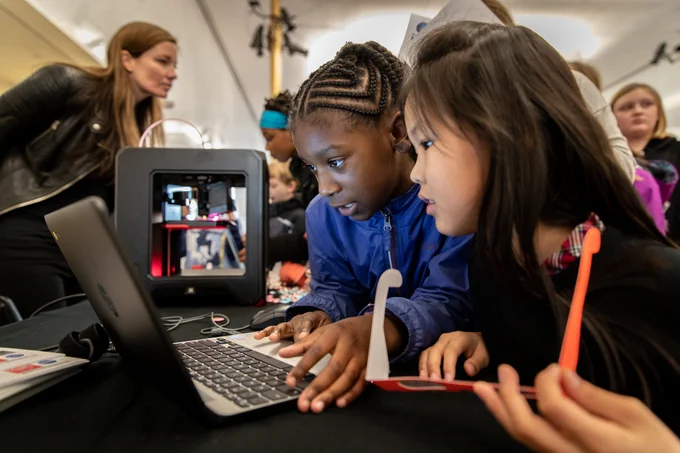 Two young students attending the ED Games Expo. The students are sitting huddled together over a laptop; one student is holding a pair of orange 3-D glasses.