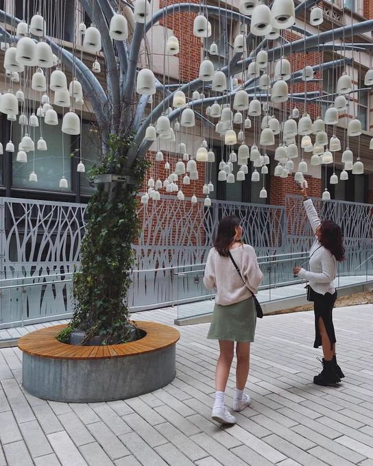 Two young women standing to the right of the Speaking Willow, a metal tree covered in ivy with bell-shaped speakers, at Planet Word. The woman to the right has her arm stretched upward to the bells.