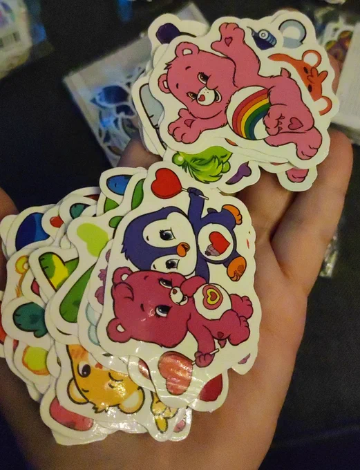 Sticker in meiner Hand. Auf den Stickern sind die Glücksbärchen abgebildet. Man sieht einen pinken Bären mit einem Herzlolli in der Hand und einem Herz auf dem Bauch. Er ist mit einem blauen Pinguin auf dem Sticker, der ebenfalls ein Herz auf dem Bauch und Lolli in der Hand hat. Daneben ist ein Sticker mit einem rosa Bärchen mit Regenbogen auf dem Bauch. Im Hintergrund sieht man noch einige Stickerpakete.