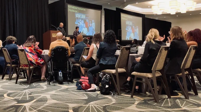 Betty Siegel on stage speaking to a conference room full of seated viewers.