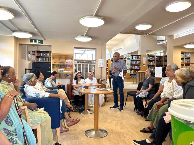 Photograph taken with Northwood Hills Library of attendees to the Gujarati Meet Up group held on Wednesdays. Attendees are sat in chairs arranged in a rectangular shape, the camera is angled at a seated level and the attendees are looking towards a gentleman who is stood to the upper right of the image talking with the group. There are two light wooden coloured round tables in the centre of the seated arrangement.